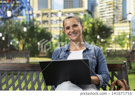 Focused woman studying content on laptop while sitting on wooden bench in urban park, with greenery and modern buildings in background. Everyday life technology use. Focused woman studying content on laptop while sitting on wooden bench in urban park, with greenery and modern buildings in background. Everyday life technology use. 141871758