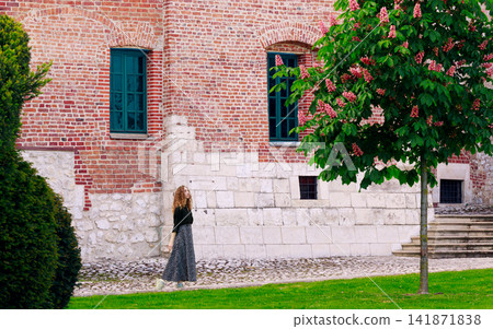charming young woman with curly hair strolls through a historic fortress courtyard adorned with blooming trees and lush greenery charming young woman with curly hair strolls through a historic fortress courtyard adorned with blooming trees and lush greenery 141871838