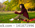A cute girl reads a book and drinks coffee on a green lawn in an autumn park. Autumn mood. A cozy place to be alone with yourself 141871996