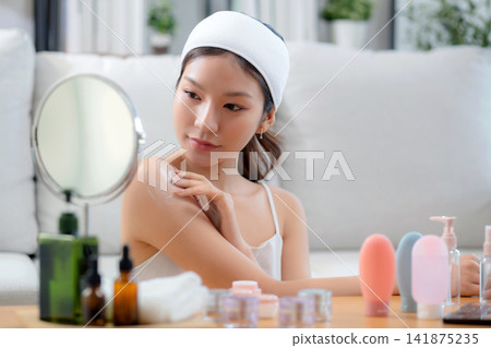 Beauty young asian woman applying cream or lotion while looking mirror in living room at home. 141875235