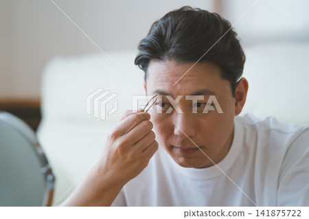 一位中年男子在桌面鏡子前用鑷子仔細地拔眉毛——男士美容程序。 141875722