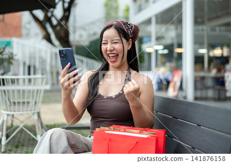 Excited asian woman shopper celebrating good news on phone with shopping bags on bench at mall plaza Excited asian woman shopper celebrating good news on phone with shopping bags on bench at mall plaza 141876158