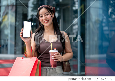 Young asian woman customer showing white screen smartphone while holding a drink and shopping bags. 141876229