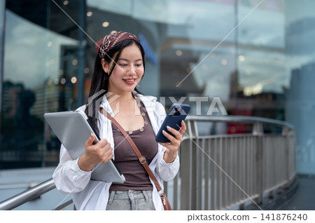 Asian Woman Looking at Smartphone while Carrying Laptop Asian Woman Looking at Smartphone while Carrying Laptop 141876240