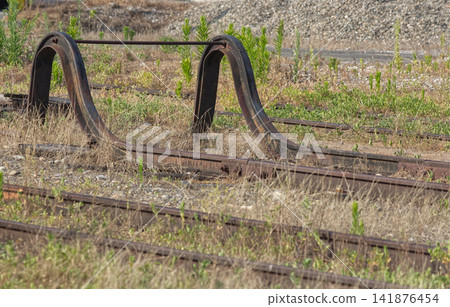 The end of the railway tracks, covered in grass, with a rusty old buffer stop and a railway landscape. The end of the railway tracks, covered in grass, with a rusty old buffer stop and a railway landscape. 141876454