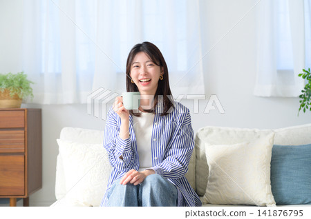 A smiling Japanese woman drinking coffee on a sofa. A smiling Japanese woman drinking coffee on a sofa. 141876795