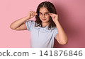 Young Girl with Glasses Posing Playfully Against a Soft Pink Background in Bright Light 141876846