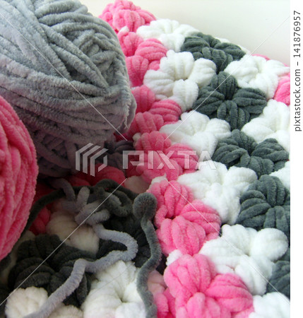 A cozy, plush handmade blanket featuring pink, white, and grey flower motifs. Balls of velvety chenille yarn rest in the foreground, highlighting a work-in-progress hobby scene. A cozy, plush handmade blanket featuring pink, white, and grey flower motifs. Balls of velvety chenille yarn rest in the foreground, highlighting a work-in-progress hobby scene. 141876957