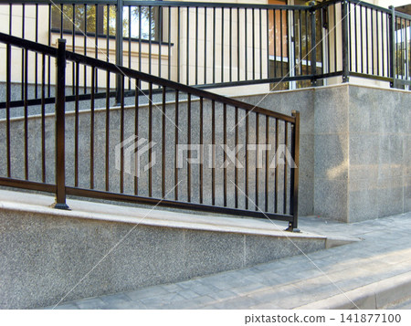 Outdoor ramp with grey stone slabs and black metal railings leading to a modern building entrance. Paved walkway in the foreground. Outdoor ramp with grey stone slabs and black metal railings leading to a modern building entrance. Paved walkway in the foreground. 141877100