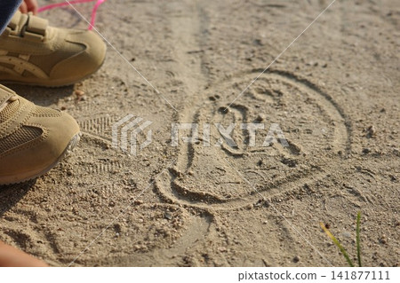 A face drawn in the sand on the park ground and a child's feet. A face drawn in the sand on the park ground and a child's feet. 141877111