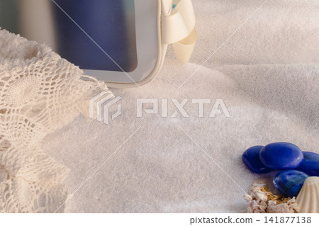 White fluffy towel creating a relaxing wellness and spa background with blue decorative stones, seashells, and crochet fabric, providing copy space for advertising or branding campaigns White fluffy towel creating a relaxing wellness and spa background with blue decorative stones, seashells, and crochet fabric, providing copy space for advertising or branding campaigns 141877138