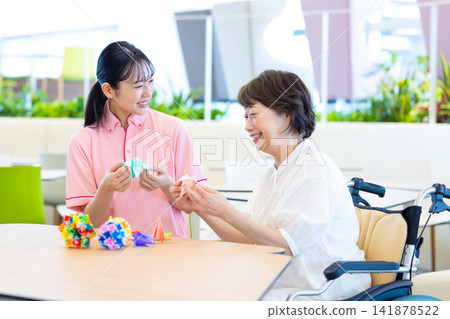 Caregiving image: Senior woman folding origami with caregiver. Photography cooperation: Denpa Gakuen Tokyo Electronics College. Caregiving image: Senior woman folding origami with caregiver. Photography cooperation: Denpa Gakuen Tokyo Electronics College. 141878522