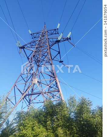 Low-angle shot of a towering steel lattice transmission pylon with high-voltage power lines and insulators, set against a clear blue sky, with green foliage at its base. Low-angle shot of a towering steel lattice transmission pylon with high-voltage power lines and insulators, set against a clear blue sky, with green foliage at its base. 141879054