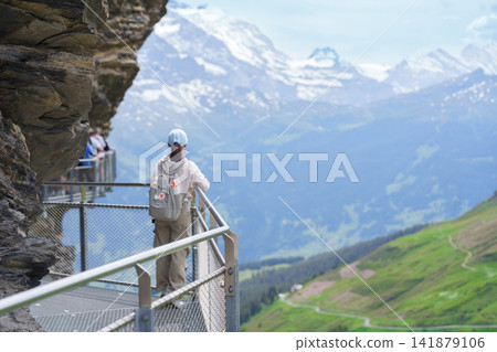 Woman Tourist waking on First Cliff Walk against Swiss Alps at Grindelwald First, Switzerland in Summer. Famous Traveler Attraction, Travel Destination and hiking trail in Europe. Top of Adventure Woman Tourist waking on First Cliff Walk against Swiss Alps at Grindelwald First, Switzerland in Summer. Famous Traveler Attraction, Travel Destination and hiking trail in Europe. Top of Adventure 141879106
