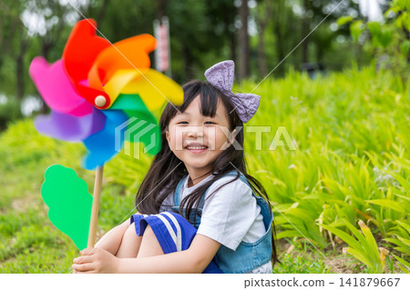 Happy Young East Asian Girl with Purple Bow Sitting in Sunny Green Meadow Holding Colorful Rainbow Pinwheel with Joyful Smile and Bright Summer Vibes. Happy Young East Asian Girl with Purple Bow Sitting in Sunny Green Meadow Holding Colorful Rainbow Pinwheel with Joyful Smile and Bright Summer Vibes. 141879667