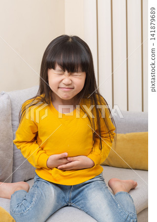 Asian little girl suffering stomach ache, sitting on sofa, painful expression, child health problem, abdominal pain concept Asian little girl suffering stomach ache, sitting on sofa, painful expression, child health problem, abdominal pain concept 141879689