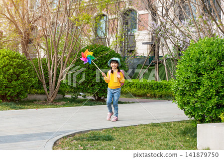 Delighted Young East Asian Girl Holding Colorful Pinwheel While Walking on Park Path in Yellow Sweater Pink Backpack and Light Blue Hat with Bright Smile. Delighted Young East Asian Girl Holding Colorful Pinwheel While Walking on Park Path in Yellow Sweater Pink Backpack and Light Blue Hat with Bright Smile. 141879750