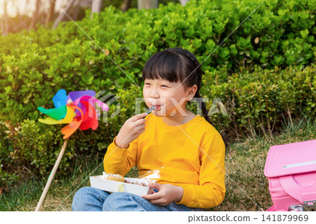 Asian little girl eating dessert in park, smiling happily, outdoor childhood lifestyle for leisure, picnic and healthy growth concept 141879969