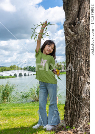 A cheerful young girl leaning against a tree trunk and raising a willow branch high in a sunny park. 141879998