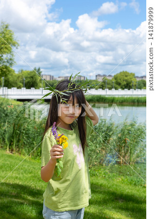 A young girl wearing a handmade willow garland and holding wildflowers smiles near a scenic riverbank. A young girl wearing a handmade willow garland and holding wildflowers smiles near a scenic riverbank. 141879999