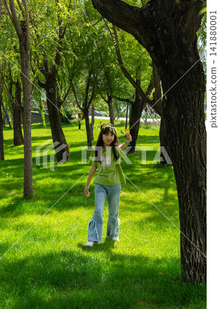 A young girl wearing a leaf crown stands amidst tall green trees on a sunlit grassy field. A young girl wearing a leaf crown stands amidst tall green trees on a sunlit grassy field. 141880001