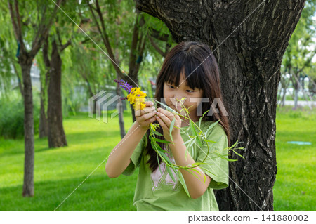 A young girl meticulously examines a bunch of colorful wildflowers while standing next to a large tree. A young girl meticulously examines a bunch of colorful wildflowers while standing next to a large tree. 141880002