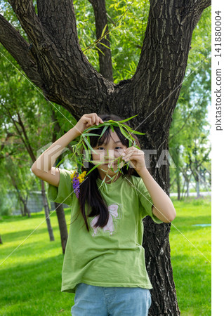 A playful young girl hiding behind a willow branch garland in front of a massive tree trunk. A playful young girl hiding behind a willow branch garland in front of a massive tree trunk. 141880004