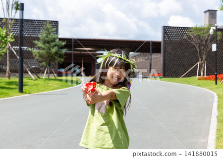 A friendly young girl extending her hand to offer a bright red flower on an empty park pathway. 141880015