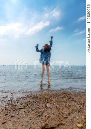 A carefree young girl in a red cap jumps high into the air on a sunny beach facing the ocean. 141880016
