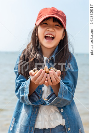 Smiling Asian little girl playing with pebbles on beach, happy child at seaside, summer vacation and outdoor leisure concept 141880017