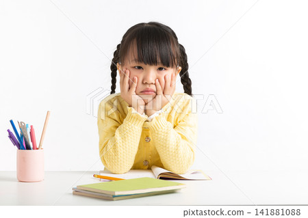 Unhappy little girl holding her chin in her hands while sitting at a desk with school books. Unhappy little girl holding her chin in her hands while sitting at a desk with school books. 141881088