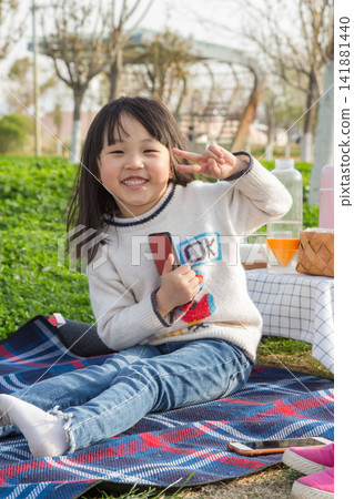 Portrait of a smiling little girl showing victory sign sitting on picnic mat in park. Portrait of a smiling little girl showing victory sign sitting on picnic mat in park. 141881440