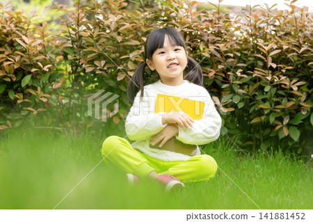 Happy little girl hugging her favorite book while sitting on green meadow, love for reading. Happy little girl hugging her favorite book while sitting on green meadow, love for reading. 141881452