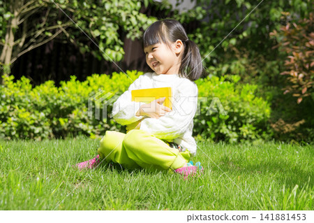 Happy little girl hugging her favorite book while sitting on green meadow, love for reading. 141881453