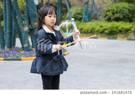 Joyful little girl holding a bubble wand and laughing with huge soap bubbles in the garden. Joyful little girl holding a bubble wand and laughing with huge soap bubbles in the garden. 141881478