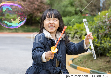 Joyful little girl holding a bubble wand and laughing with huge soap bubbles in the garden. 141881482