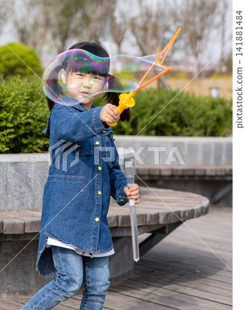 Joyful little girl holding a bubble wand and laughing with huge soap bubbles in the garden. Joyful little girl holding a bubble wand and laughing with huge soap bubbles in the garden. 141881484