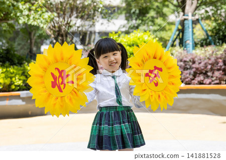 Joyful little girl in school uniform holding large paper sunflowers during a campus activity.The text on the flower reads: "Happiness." Joyful little girl in school uniform holding large paper sunflowers during a campus activity.The text on the flower reads: "Happiness." 141881528