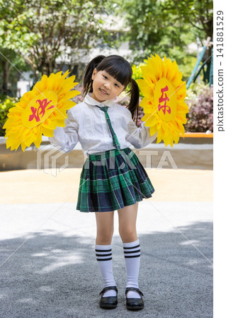 Joyful little girl in school uniform holding large paper sunflowers during a campus activity.The text on the flower reads: "Happiness." 141881529