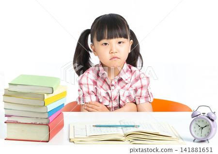 Unhappy student sitting with arms crossed behind a desk with school books and clock. Unhappy student sitting with arms crossed behind a desk with school books and clock. 141881651