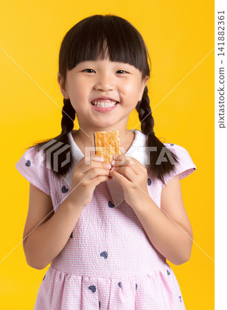 Happy smiling girl holding a crispy wafer biscuit with both hands on yellow background Happy smiling girl holding a crispy wafer biscuit with both hands on yellow background 141882361
