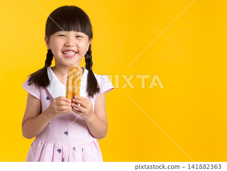 Smiling little girl holding a long pastry puff snack against a vibrant yellow background Smiling little girl holding a long pastry puff snack against a vibrant yellow background 141882363