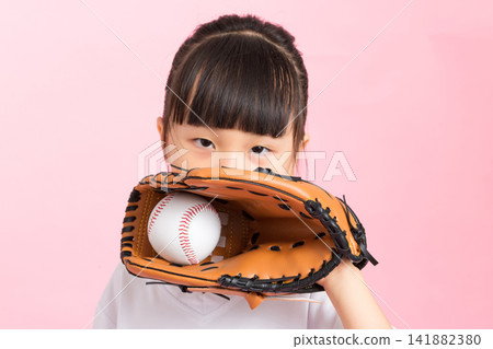 Close up portrait of a girl peeking behind a baseball leather glove holding a ball on pink background. Close up portrait of a girl peeking behind a baseball leather glove holding a ball on pink background. 141882380