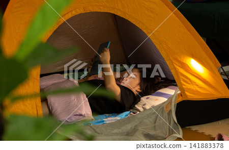 Little girl lying in a cozy indoor tent at night, using a tablet or phone, glamping and home adventure concept. 141883378