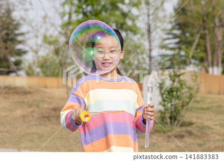 Portrait of a smiling young girl in a colorful sweater playing with a large soap bubble outdoors. Portrait of a smiling young girl in a colorful sweater playing with a large soap bubble outdoors. 141883383