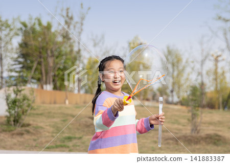 Portrait of a smiling young girl in a colorful sweater playing with a large soap bubble outdoors. Portrait of a smiling young girl in a colorful sweater playing with a large soap bubble outdoors. 141883387