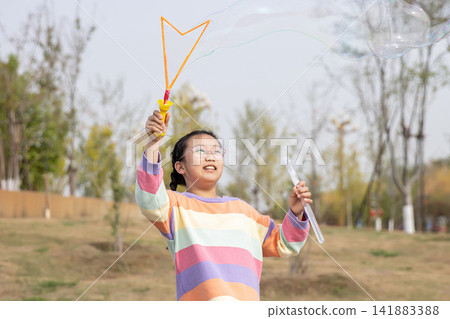 Portrait of a smiling young girl in a colorful sweater playing with a large soap bubble outdoors. Portrait of a smiling young girl in a colorful sweater playing with a large soap bubble outdoors. 141883388