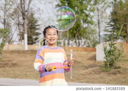 Portrait of a smiling young girl in a colorful sweater playing with a large soap bubble outdoors. Portrait of a smiling young girl in a colorful sweater playing with a large soap bubble outdoors. 141883389