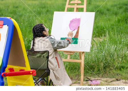 Young girl student painting on a canvas in a vast green meadow during art class. Young girl student painting on a canvas in a vast green meadow during art class. 141883405