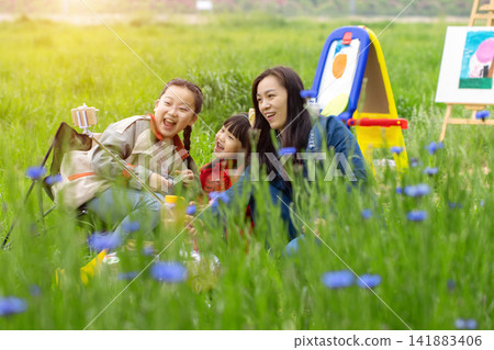 Happy Asian family with mother and two daughters taking a selfie while painting in a meadow field. Happy Asian family with mother and two daughters taking a selfie while painting in a meadow field. 141883406